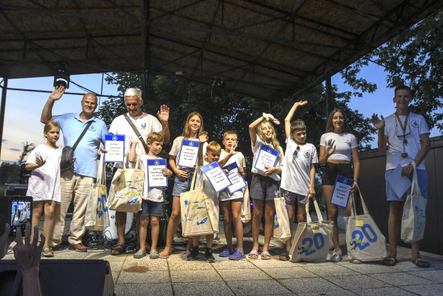 A group of children and adults stand on a stage holding certificates and Danube Day tote bags, smiling and waving during an award ceremony outdoors.