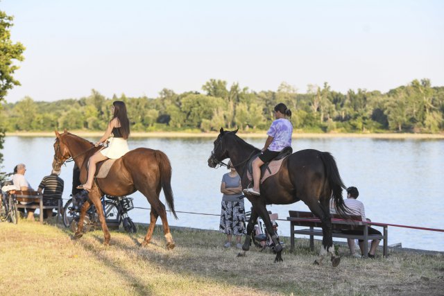 Two people ride horses along a grassy riverbank while others sit on benches enjoying the view across the calm water.