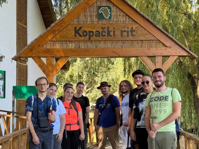 Several people gather outside under a wooden sign. 