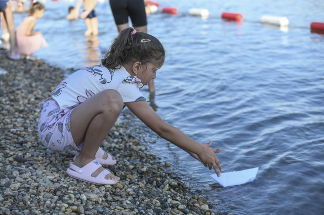 A young girl crouches by the water’s edge on a pebbled beach, gently placing a small paper boat into the water.