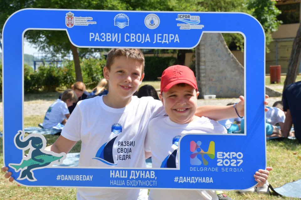 Two smiling children wearing white event shirts pose together outdoors, holding a large blue photo frame with logos and Serbian text promoting the Danube and EXPO 2027.