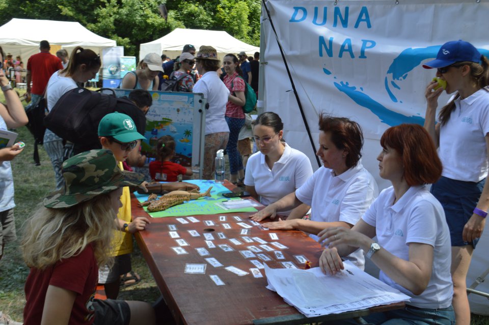 Children look at a table with various cards related to Danube Day, with adults sitting behind the table in white t-shirts.