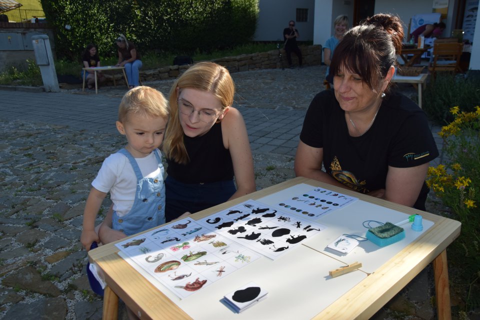 A young child and two adults sit together at an outdoor table examining a collection of small educational objects arranged on display boards.